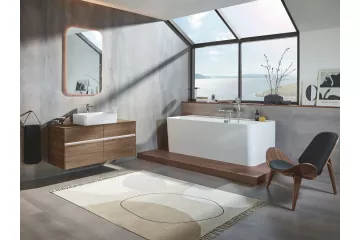 Elegancka łazienka Villeroy & Boch z dużą wolnostojącą wanną i drewnianą szafką pod umywalkę. Przestronne wnętrze z panoramicznym widokiem na morze, designerskim fotelem i minimalistycznymi dekoracjami.